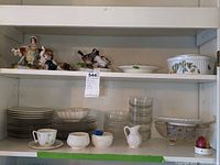 Wide shelf view showing assorted porcelain plates, bowls, cups, glass bowls, and animal figurines including geese and tiger figurine.