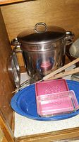 Photo showing stainless steel Crock-Pot with lid, large metal pot, blue speckled baking platter, plastic storage containers, and wooden utensil.