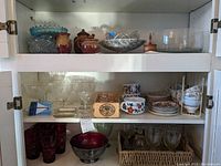 Overall view of cabinet shelf with mixed glassware, pottery, wooden box, and tea sets arranged on three shelves.