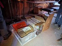 Three boxes on the floor including a clear plastic storage bin and cardboard boxes containing glassware