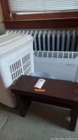 Photo showing white plastic laundry hamper, small brown wooden bench, and white Lakewood heater grouped together by a window.