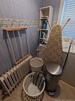 Photo showing the ironing board with tropical palm tree print leaning against the wall near radiators. White plastic laundry baskets and a metal bucket hamper on the floor along with a stainless steel trash can with foot pedal. Wooden drying rack partially visible.