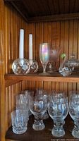 Top shelf showing faceted glass candle holders holding white taper candles, iridescent champagne flute, frosted goblet, and one glass swan along with glass bowls below.