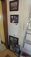 Framed pictures arranged on a wall and floor next to a ladder, including image of children and historic buildings