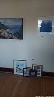 Full view showing multiple framed artworks on and near a wall: large coastal oil painting, blue abstract face print, lighthouse painting, and smaller framed pieces on the floor.