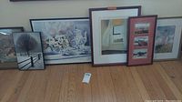 Photo of six framed artworks on the floor including floral still life, winter tree scene, woman in field, airplane photos, and landscape.
