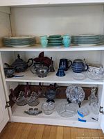 Full view of cabinet shelves with stacked Wedgewood dinner and dessert plates on top shelf; silver plated teapot set, blue mugs, and other items on middle shelf; cut glass and silver plated items on bottom shelf.