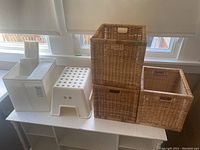 Three wicker baskets and a plastic step stool displayed on a white surface near windows.