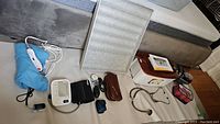 Overview of health and beauty devices on soft surface including blood pressure monitors, pulse oximeter, heating pad, paraffin bath machine, hair curlers, stethoscope, and small bags.
