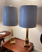 Two table lamps side by side on wooden surface showing white plastic shades and gold-tone painted wood fluted column bases with classical motifs.