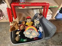 Full view of the gray tote bin containing vintage plush toys including Bugs Bunny, Curious George, Raggedy Ann, a wooden rainbow clock, and a red plastic skating trainer frame behind the bin.