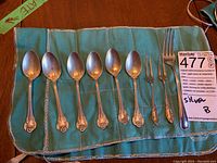 Photo showing six tea spoons and three forks laid out on a teal cloth.