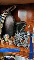 Shelf display with large metal German folk art cow bell on decorative fabric belt, small souvenir keychain bells painted with floral designs, metal warrior and animal figurines, some broken.