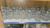 Wide shot showing 18 etched glass German beer steins arranged in a row on a shelf, all with pewter lids.