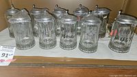 Ten etched glass steins with pewter lids arranged on a shelf, showing the full set and etched details.