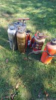 Photo showing 8 vintage metal fire extinguishers and one metal sprayer standing on grass outdoors, various sizes and finishes with visible wear and dirt.