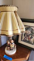 Photo showing the lamp with cream-colored pleated fabric shade accented with gold trim and hanging tassel. Lamp base shows two figurines of children in detailed attire on a round white base. Slight damage visible on top edge of shade. 3D framed art with image of children in raincoats behind glass is visible in background.