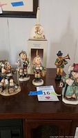 Full view of three vintage Hummel figurines, the 1985 Hummel annual bell on box, Friedel figure, and wooden horn player arranged on wooden surface
