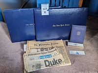 Wide view of all items showing blue binders, newspapers, pen box, and smaller pamphlet.