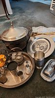 Overview of plated servingware and pewter collectible items on carpeted floor including chafing dish, trays, small containers and plates.