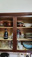 Wooden cabinet shelves filled with various vintage kitchenware including glazed bowls, clear glass cups, ramekins, stained sauce cups and a black manual chopper.