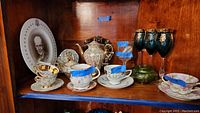 Overall view of the tea set including cups, saucers, teapot, green lidded pot, Eisenhower plate, and blue gold-accented stemware on a wooden shelf