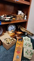 Wooden shelf holding various items including wooden carved ducks, two Asian style trays stacked, brass candle holders, decorative plates, rattan box, ceramic items, and wall shelf visible