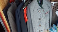 Multiple jackets and suit pieces hanging in a closet, including grey jacket with embroidery and decorative buttons, and dark colored jackets.