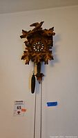 Full view of the cuckoo clock hanging on a wall, showing the carved leaves, birds, clock face, and hanging weights.