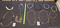 Eight various necklaces laid out flat on a black surface including different styles and materials like cord, beads, chains.