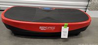 Side angle view of the Body Express Curve vibration plate showing the black rubberized top surface, red edging, and the brand logo on the front side.