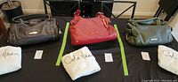 All three Cole Haan handbags in brown, red, and olive green arranged side by side on a table with their white fabric storage dust bags below each handbag.