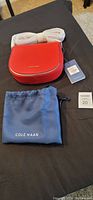 Front view of new red Cole Haan handbag with tags and dust bag on black surface.