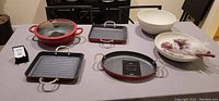 Overview of Curtis Stone cookware including red multi-pan with glass lid, gray grill pan, two white melamine bowls with utensils set on a table