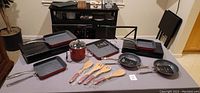 Full set view of Curtis Stone cookware on table including frypans, square grill pan, pot, wooden spoons, and oven gloves