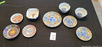 Nine Japanese decorative porcelain items, six plates and three bowls, displayed on black cloth.