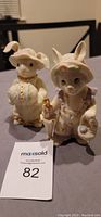 Two of the four Lenox Easter rabbit figurines displayed side by side on a gray surface. Both rabbits are in vintage-style outfits, one holding an umbrella, the other with a round decorated item.