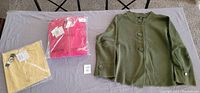 Three sweaters displayed on a table: an olive green sweater topper with large buttons, a yellow sweater, and a pink zip-up sweater in plastic packaging with tags attached.