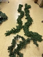 Photo showing two long green artificial Christmas garlands laid flat on a tiled floor, showing full length and needle details.