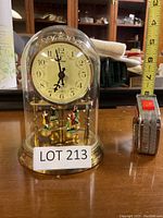Front view of anniversary clock inside glass dome, showing clock face with floral accents, black ornate hands, and four colorful figurines at the base.