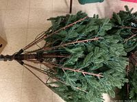 Photo shows the artificial Christmas tree folded with visible brown branch stems and green plastic needles. Tree is bound together without stand.