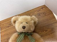 Full view of Barrington Bear showing light brown fur and green bow around neck, positioned on a wooden surface with white sidewalls in background.