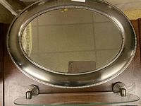 Oval mirror with brushed silver metal frame and beaded border, reflection shows ceiling tiles and phone selfie