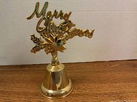 Gold-tone metal bell approximately 7 inches tall with 'Merry Christmas' script topper and poinsettia decoration on top, photographed from front.