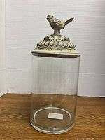 Full view of clear glass container with ornate covered lid topped by bird figurine shown against white background on wooden surface.