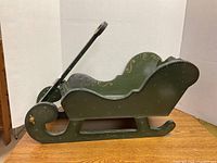 Side view of the green painted wooden sleigh with gold swirl designs and a handle.