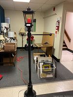 Tall black vintage style street lamp with square lantern top and red LED candle inside, shown in a basement room with other items and orange extension cord on floor.