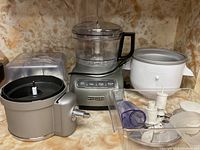 Photo showing KitchenAid food processor base with multiple attachments and parts on countertop with marbled background