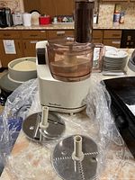 Full view of Moulinex food processor with attached translucent brown bowl and lid, two slicing/shredding discs on plastic sheet in front.