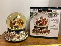 Musical water globe with Santa and reindeer inside glass globe, sitting on a decorated revolving base with village and animal details, alongside its original box.